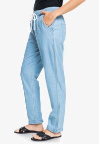 Pantaloni di tessuto morbido blu chiaro con elastico in vita e cordoncino bianco. Presentano tasche laterali e una vestibilità comoda. Indossati con sandali neri.