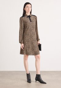 Tierdruckkleid in Beige mit schwarzen Punkten, ausgestattet mit einem schwarzen Kragen und langen Puffärmeln. Kombiniert mit schwarzen Ankle Boots und einer Clutch.
