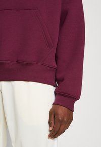 Mörkhyad person iklädd en vinröd huva med en framficka och off-white byxor, som visar nedre delen av kroppen och höger hand.