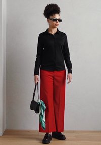Camicia nera con bottoni, pantaloni larghi rossi, mocassini neri, piccola borsa nera con dettaglio di sciarpa turchese, occhiali da sole, capelli ricci.
