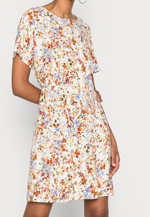 Kvinde iført hvid kjole med korte ærmer med flerfarvet blomsterprint og et tyndt bælte i taljen, stående mod en ensfarvet baggrund.
