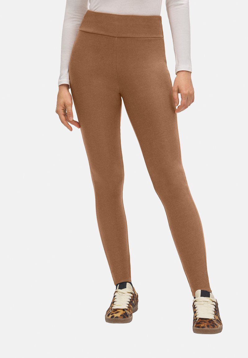 Leggings marrones con una cintura alta, hechos de material suave, diseñados con una textura lisa; combinados con zapatillas de deporte marrones y negras con un patrón.