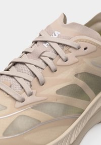 Μπεζ αθλητικό παπούτσι HOKA με άνω μέρος από δίχτυ, παχιά επενδεδυμένη σόλα και επίπεδα μπεζ κορδόνια δεμένα σε σταυρωτό μοτίβο.