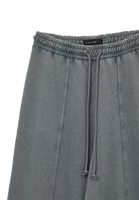 Pantalon de survêtement gris avec taille élastique et cordon de serrage. Dispose de détails de couture sur les côtés et d'une texture lisse et douce. Étiquette de marque visible.