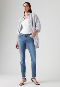 Fehér ujjatlan top csíkos ing alatt, kék skinny farmerral és fekete magassarkú szandállal párosítva. Az ingnek felgyűrt ujjai és laza szabása van.