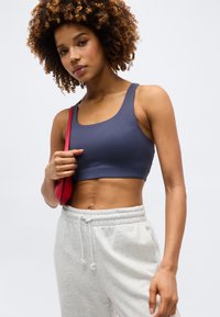 Γυναίκα με σγουρά μαλλιά που φοράει μπλε σπορ τοπ και ανοιχτό γκρι φόρμα, κρατώντας μια μικρή κόκκινη τσάντα ώμου.