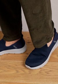 Marinefarbene Slipper mit strukturiertem Mesh-Obermaterial und weißer Gummisohle, kombiniert mit dunkelgrünen Cordhosen. Darunter ein Holzfußboden.