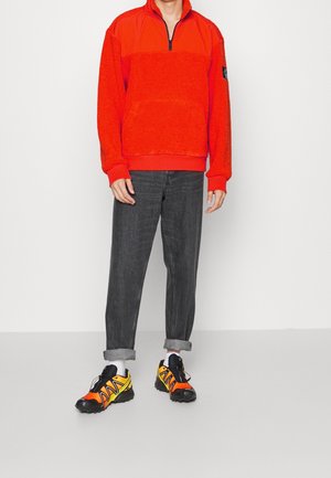 Roter Fleecepullover mit Reißverschluss und durchgehendem Futter, mit einem soliden unteren Bereich, kombiniert mit dunklen Jeanshosen und schwarzen/orangen Laufschuhen.