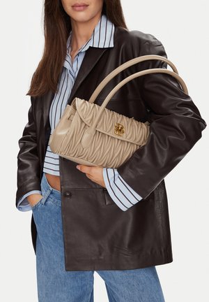 Žena v tmavě hnědém koženém saku, modré pruhované košili a džínách držící béžovou prošívanou koženou kabelku se zlatým logem.