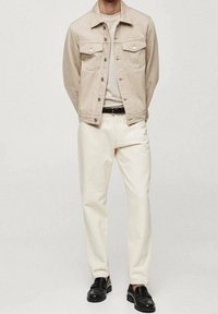 Chaqueta de mezclilla beige con dos bolsillos en el pecho, llevada sobre una camiseta clara. Los pantalones color crema y los mocasines negros completan el atuendo.