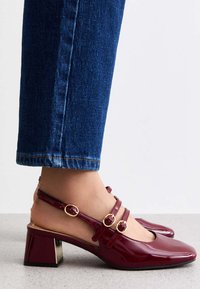 Vinröda lackläders slingback-skor med fyrkantiga tår, tre dekorativa spännband och en blockklack med en guldaccent.