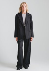 Czarna, dopasowana marynarka z wyciętymi klapami, zapinana na jeden guzik, z przednią kieszenią, zestawiona z szerokimi, czarnymi spodniami i teksturowanymi butami.