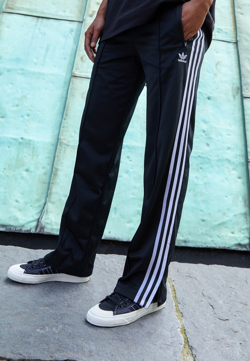 Pantalons de sport noirs avec trois bandes blanches sur les côtés. Associés à des baskets noires et blanches avec une finition en toile et des lacets plats.