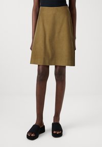Falda A-line de pana verde oliva con acanaladuras verticales, hasta la mitad del muslo. Combinada con sandalias deslizantes negras que tienen una suela gruesa.