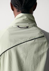 Veste vert clair texturée avec un col montant, dotée d'une ligne d'accent noir et d'un détail en loop tressé au dos du col.