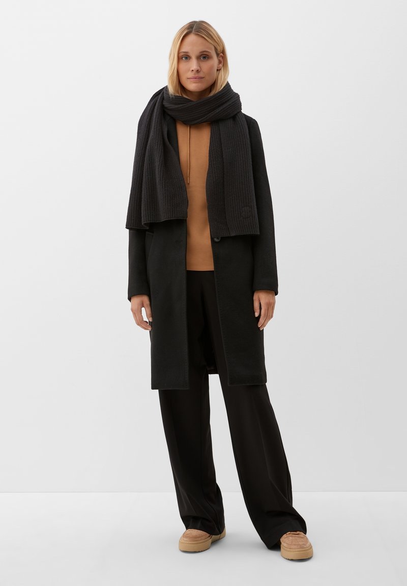 Cappotto lungo nero sopra un top sabbia, abbinato a pantaloni larghi neri e sciarpa nera a coste. Gli accessori includono scarpe con tacco chiaro.