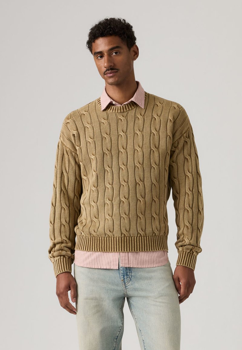 Man wearing a tan cable-knit sweater over a pink striped collared shirt and light blue jeans, standing against a plain background.