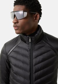 Sort pufferjakke med chevron-quiltning, høj krave og lynlåsdetalje. Reflekterende, oversized solbriller med sort stel.