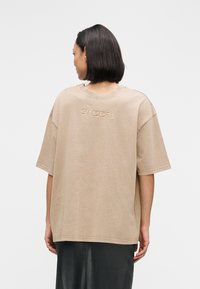 T-shirt en coton beige surdimensionné, manches courtes, encolure ronde, avec texte brodé contrastant au dos. Coupe fluide et décontractée.