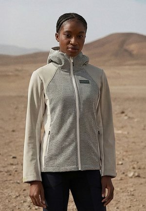 Donna in piedi in un paesaggio desertico, indossando una giacca con cappuccio a zip di colore grigio chiaro e beige e pantaloni neri.