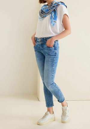 Personne portant un t-shirt blanc, une écharpe bleue à motifs, un jean bleu clair avec une taille à double bouton, et des baskets blanches, debout les mains dans les poches.