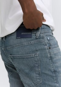 Eine Person trägt hellblaue Jeans mit einer gesteppten Gesäßtasche und hält ein dunkelblaues Etikett mit der Aufschrift "Denim Design Studio Northern Aesthetics".