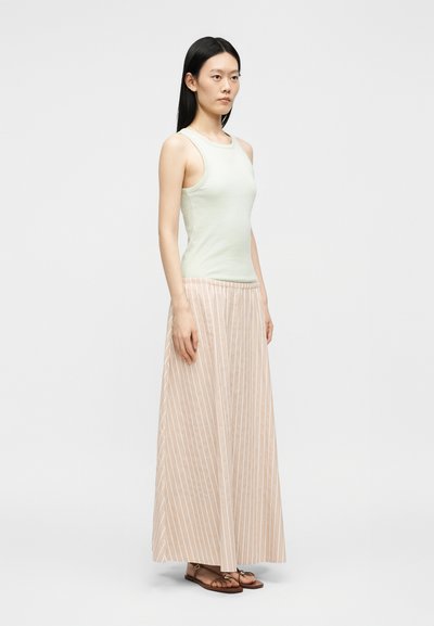Jeune femme debout portant un haut sans manches vert clair, une longue jupe beige et blanche à rayures verticales et des sandales marron sur un fond uni.