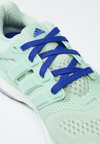 Jasnozielony but sportowy z oddychającą siateczkową cholewką, fioletowymi sznurowadłami i teksturowaną podeszwą. Posiada oznaczenia marki Adidas oraz wzmocnioną strukturę wspierającą.