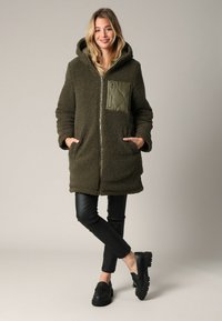 Olijfgroen fleece jas met een capuchon, voorzien van een gestikt olijfgroen zakje en twee zijzakken. Zwarte slim fit broek en schoenen maken de outfit compleet.