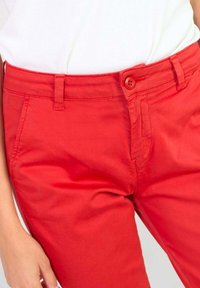 Pantalons en coton rouge avec une coupe ajustée, dotés d'un bouton de fermeture, de poches latérales et de détails de couture visibles. Texture lisse.