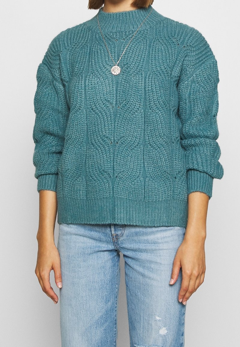 maglione a maglia turchese con motivo a trecce, polsini e orlo a costine, abbinato a jeans chiari strappati. Collana d'argento visibile.