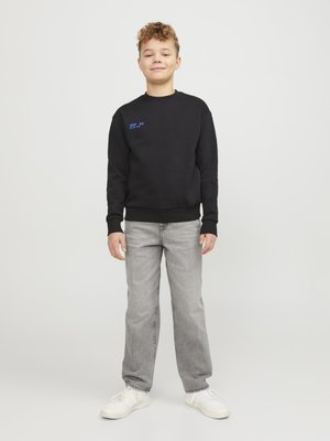 Αγόρι που στέκεται, φοράει μαύρο φούτερ με μπλε κείμενο, ανοιχτό γκρι τζιν και λευκά αθλητικά παπούτσια, μπροστά σε απλό ανοιχτόχρωμο φόντο.