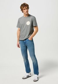 T-shirt grigio in cotone con logo circolare color crema, jeans in denim blu e scarpe da ginnastica in bianco e nero con accenti grigi. Outfit semplice e casual.
