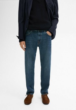 Man draagt een donkerblauwe blazer, zwarte trui, blauwe jeans en bruine suède schoenen, staat met één hand in de zak.