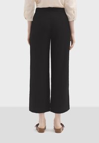 Pantalon noir à large jambe en tissu texturé. Doté d'une taille haute, il arrive jusqu'à la cheville, associé à des sandales à plateforme marron.