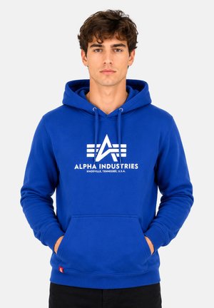 Jonge man draagt een blauwe hoodie met wit Alpha Industries-logo en tekst, handen in de voorkant zak, staand tegen een effen achtergrond.
