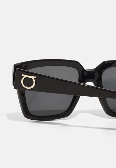 Occhiali da sole neri con una montatura spessa e angolare. Le lenti scure presentano un logo circolare dorato sul lato. Materiale in plastica lucida.