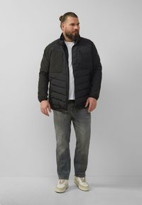 Veste noire rembourrée avec un col montant, dotée de deux grandes poches poitrine et d'une fermeture éclair à l'avant. Portée sur une chemise blanche et un jean gris.