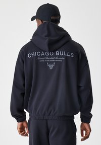 Sudadera con capucha negra que presenta "Chicago Bulls" y el logotipo en texto blanco en la parte posterior. Hecha de tela suave con puños y dobladillo de canalé.