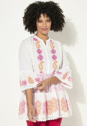 Tunique blanche avec broderie florale colorée en rose et orange, manches évasées, et dentelle au niveau de l'ourlet. Fermeture à boutons sur le devant.