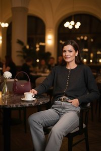 Frau in dunklem Pullover und Jeans sitzt an einem Marmortisch mit Kaffeetasse und burgunderroter Handtasche in einem sanft beleuchteten Café mit Bogenfenstern.