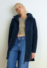 Granatowy polarowy płaszcz z zamkiem błyskawicznym, przednią kieszenią i kapturem. Nosi go się na jasny crop top i dżinsy z wysokim stanem w kolorze niebieskim. Teksturowana tkanina.