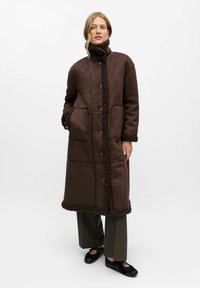 Manteau long marron avec un col et un ourlet texturés, doté de grands boutons, de poches avant et d'une finition lisse. Associé à un pantalon gris.