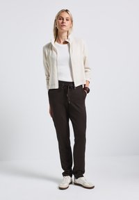 Helle beige Bomberjacke über einem weißen T-Shirt, kombiniert mit dunklen Kordelzug-Hosen und cremefarbenen Sneakers. Glattes Material mit minimalen Details.