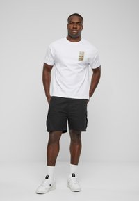 Hvid bomulds t-shirt med et front patch-design, parret med sorte cargo shorts. Modellen bærer hvide sneakers med sorte accenter og hvide sokker.