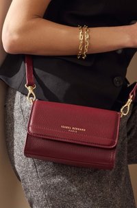 Sac en cuir bourgogne à bandoulière avec une finition texturée, accessoires en or et fermeture à rabat. Présente le nom de la marque embossé sur le devant.