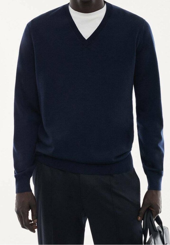 Homme portant un pull à col en V bleu marine sur une chemise blanche, tenant un sac en cuir noir, montré du cou aux hanches.