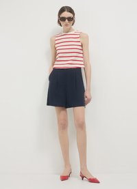 Haut sans manches blanc à rayures rouges, associé à un short taille haute bleu marine. Le mannequin porte des lunettes de soleil noires et des escarpins rouges à bout pointu.