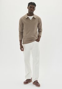 Hombre vestido con una camisa de manga larga beige, pantalones blancos y zapatos marrones, de pie con una mano en el bolsillo frente a un fondo blanco.