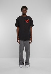Černé oversized tričko s červeným grafickým textem, spárované se šedými kalhotami s širokým střihem a bílými teniskami. Jednoduché pozadí zvýrazňuje zaměření na oblečení.